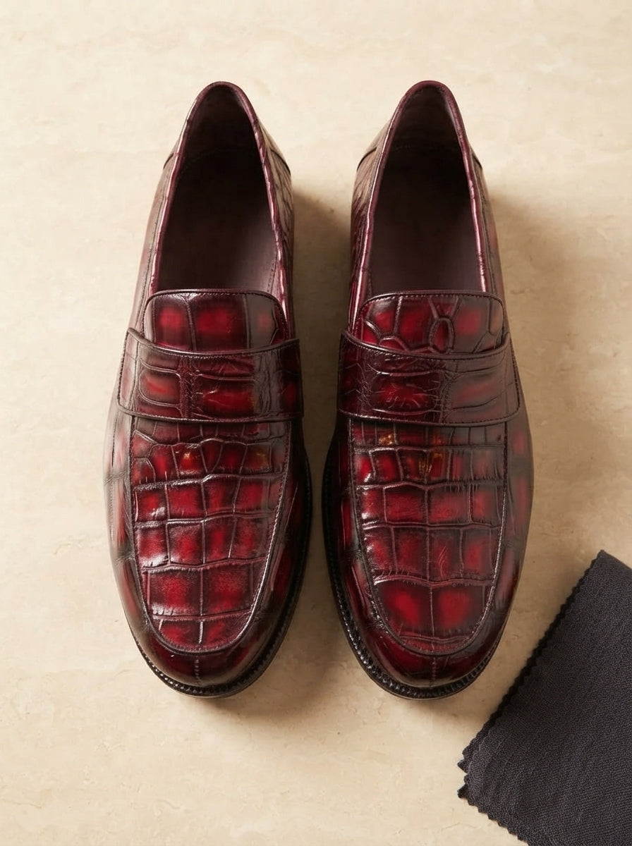 Men's red crocodile-embossed leather loafers displayed on a cream-colored stone surface with a charcoal linen accent.